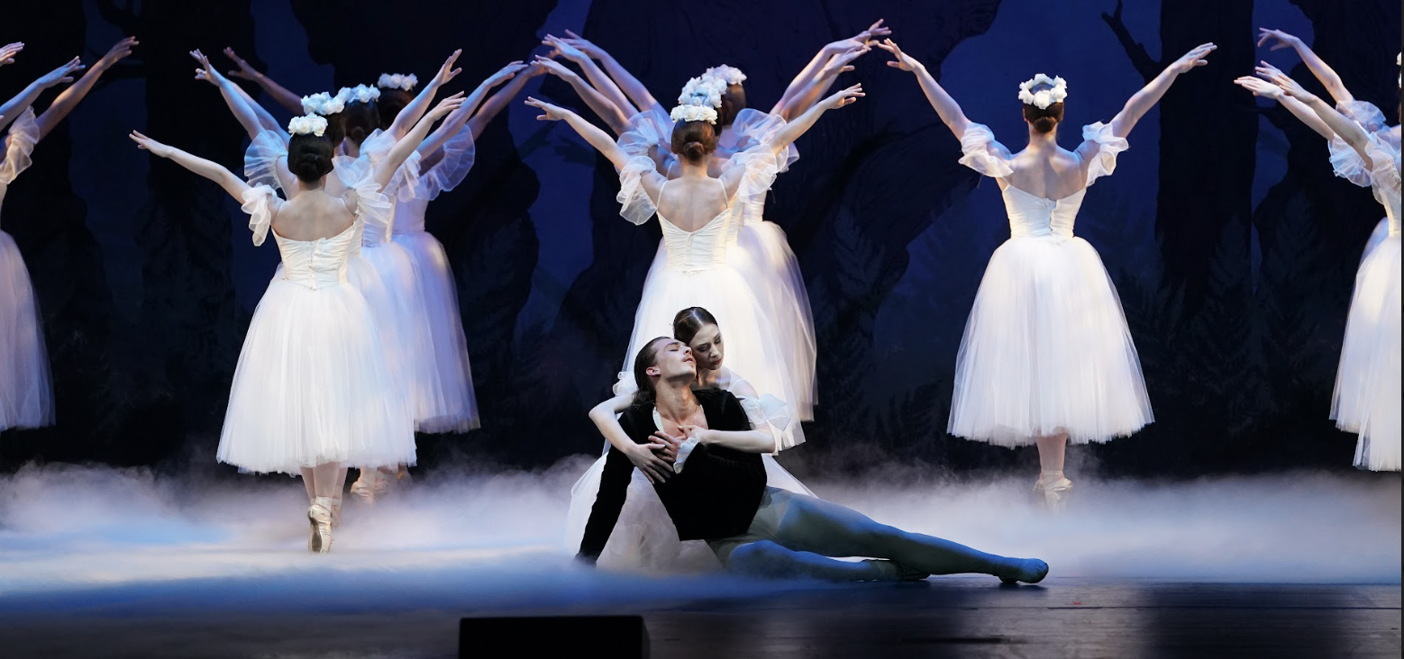 Two dancers embraced on stage with dancers in white facing backwards creating a dramatic performance.